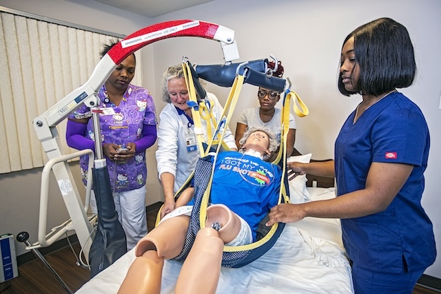 Medical training scene with nurses and an instructor guiding a patient manikin on a hospital bed using a cervical immobilizer and traction equipment for a simulation exercise.