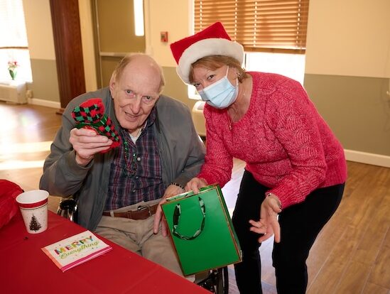 Elderly man in a wheelchair holds a knitted red-and-green item while a smiling caregiver in a Santa hat hands him a green gift bag during a Christmas celebration.