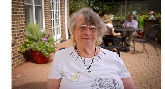Portrait of an elderly woman with short gray hair and glasses, sitting at an outdoor patio with flowers and a brick wall behind her.