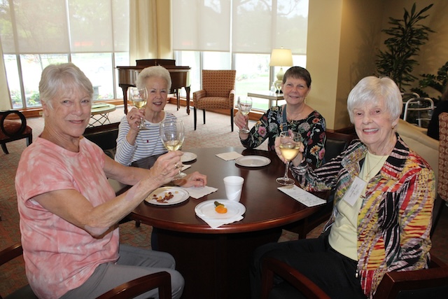 Four seniors at a round table in a bright lounge, toasting with wine glasses; plates of dessert and napkins on the table.
