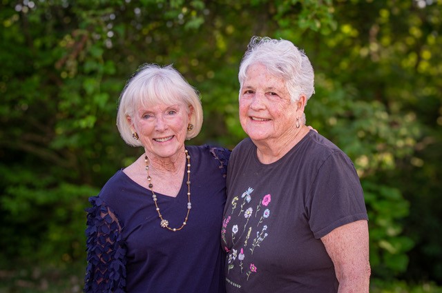 Two smiling elderly women stand close together outdoors, with greenery behind them; one wears a navy top and beaded necklace, the other a gray floral T-shirt.