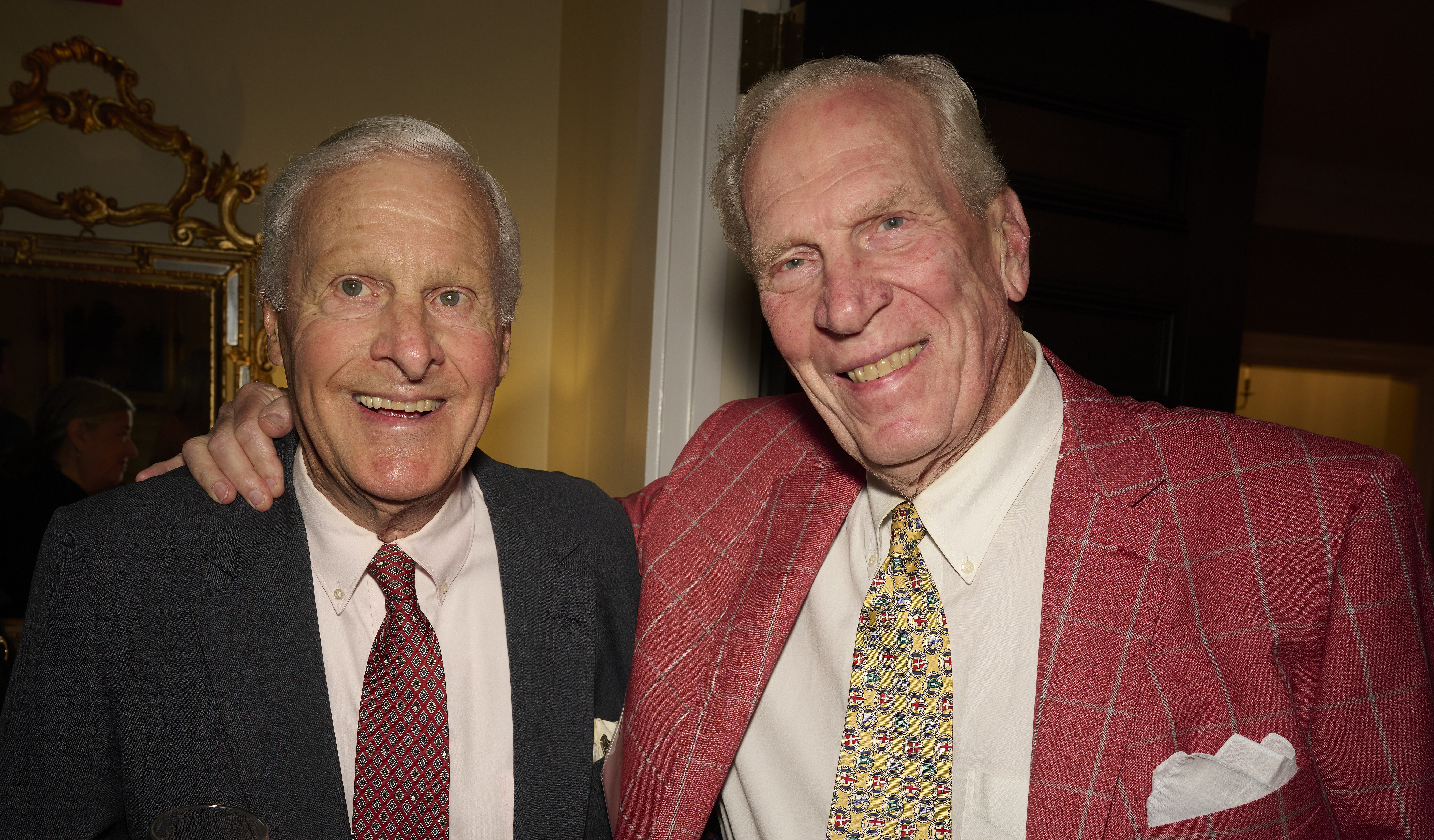 Two smiling older men in suits posing together at a social event, one wearing a dark blazer and red patterned tie, the other in a red checkered blazer with a light-colored tie and pocket square.