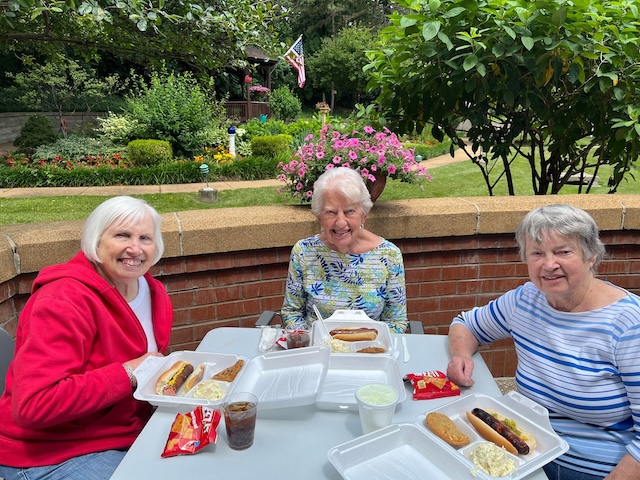 Three smiling women sit at an outdoor table with hot dogs and sides, enjoying a meal in a garden setting.