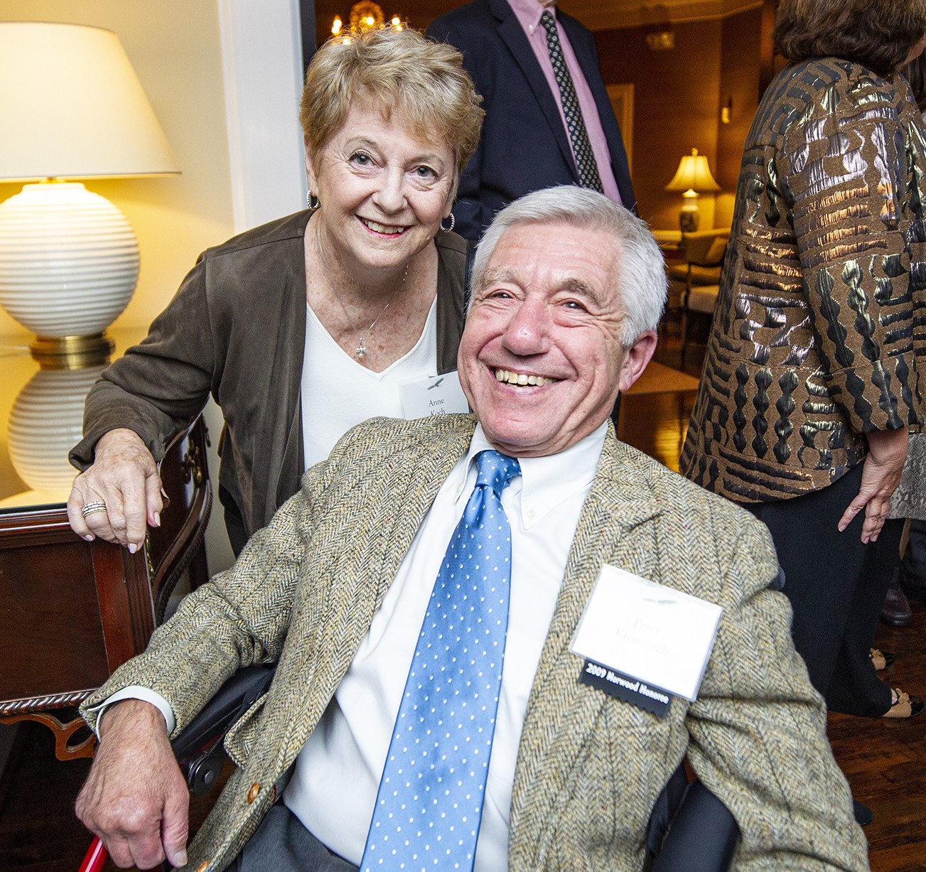 Smiling woman leaning over a man in a wheelchair at a social event; he wears a tweed jacket and blue polka-dot tie.