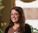 Smiling woman with wavy brown hair wearing a black cardigan, indoors with plants in the background and a flower pendant necklace.