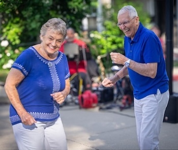 Smiling elderly couple dancing outdoors, a woman in blue top and white pants and a man in a blue polo and light trousers.