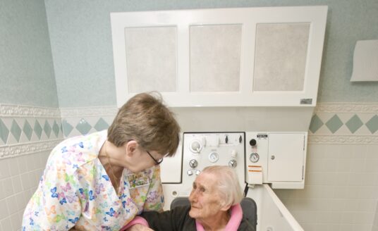 Healthcare worker in butterfly-patterned scrubs helps an elderly woman sit.