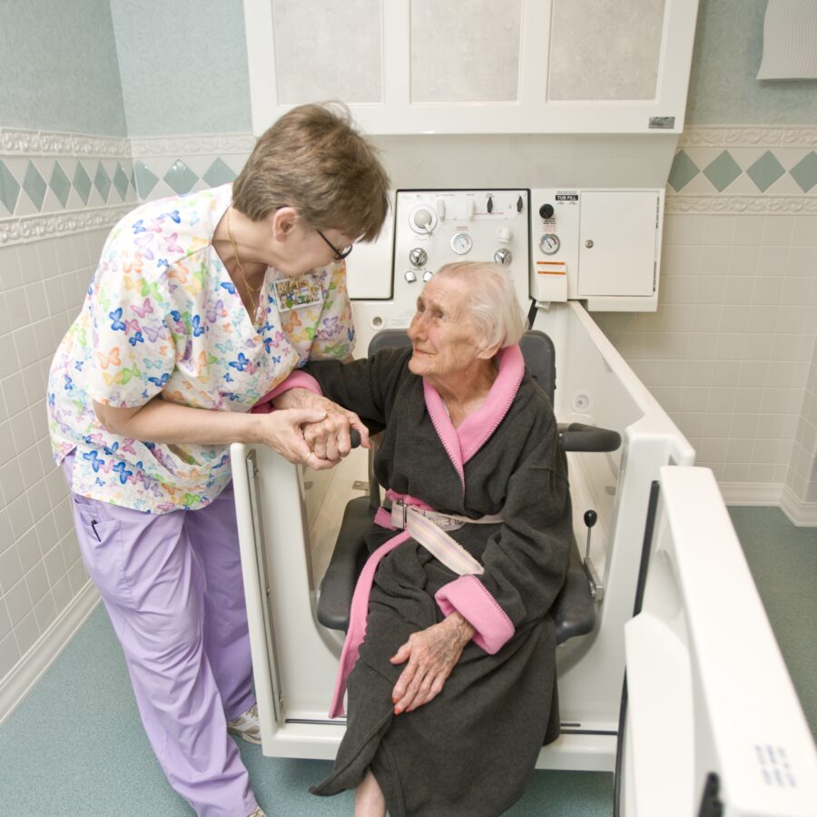 Healthcare worker in butterfly-patterned scrubs helps an elderly woman sit.