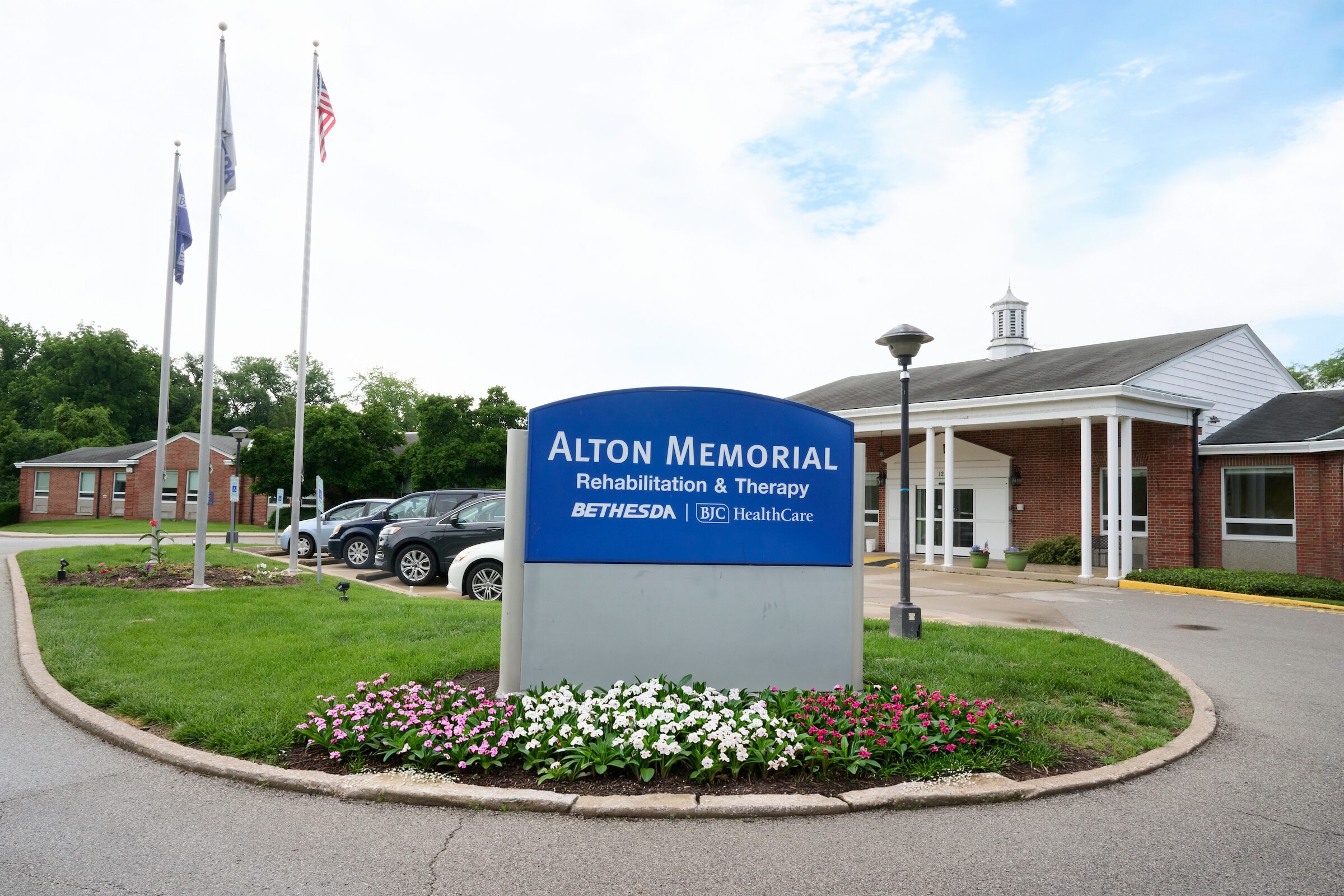 Blue sign reading 'Alton Memorial ... Rehabilitation & Therapy' in front of a brick medical building with flagpoles and a manicured flower bed nearby