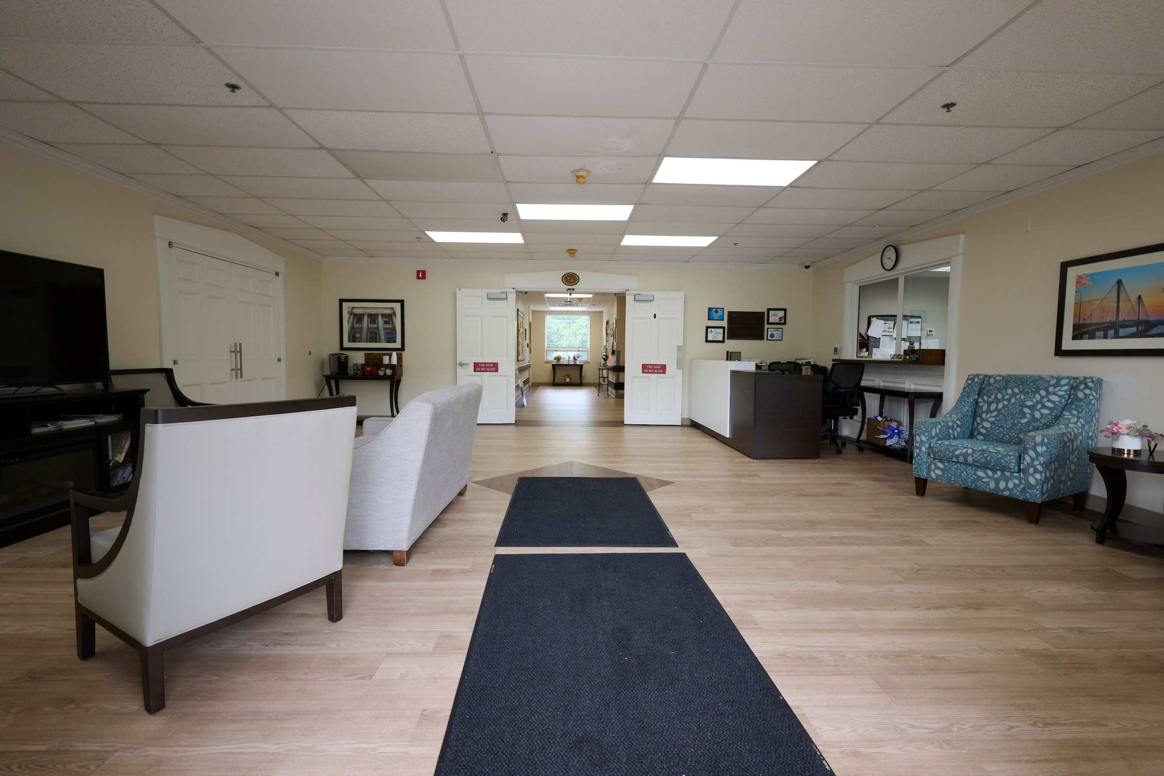 Spacious waiting room with beige walls, white doors, framed art, and blue patterned armchairs; dark blue rugs on light wood floor.