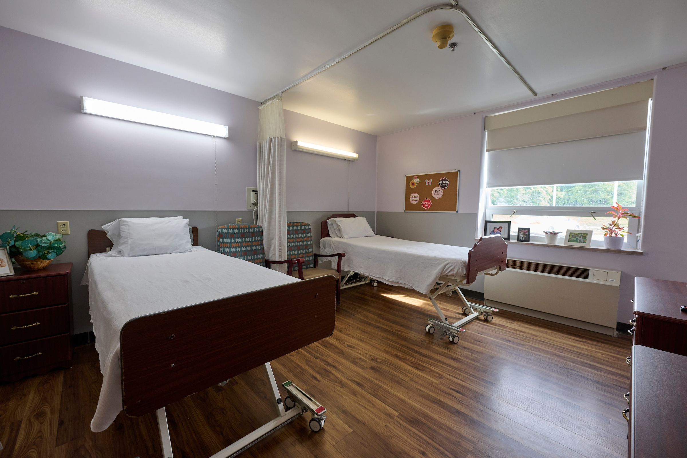 Hospital patient room with two beds, privacy curtain, and large window letting in natural light.