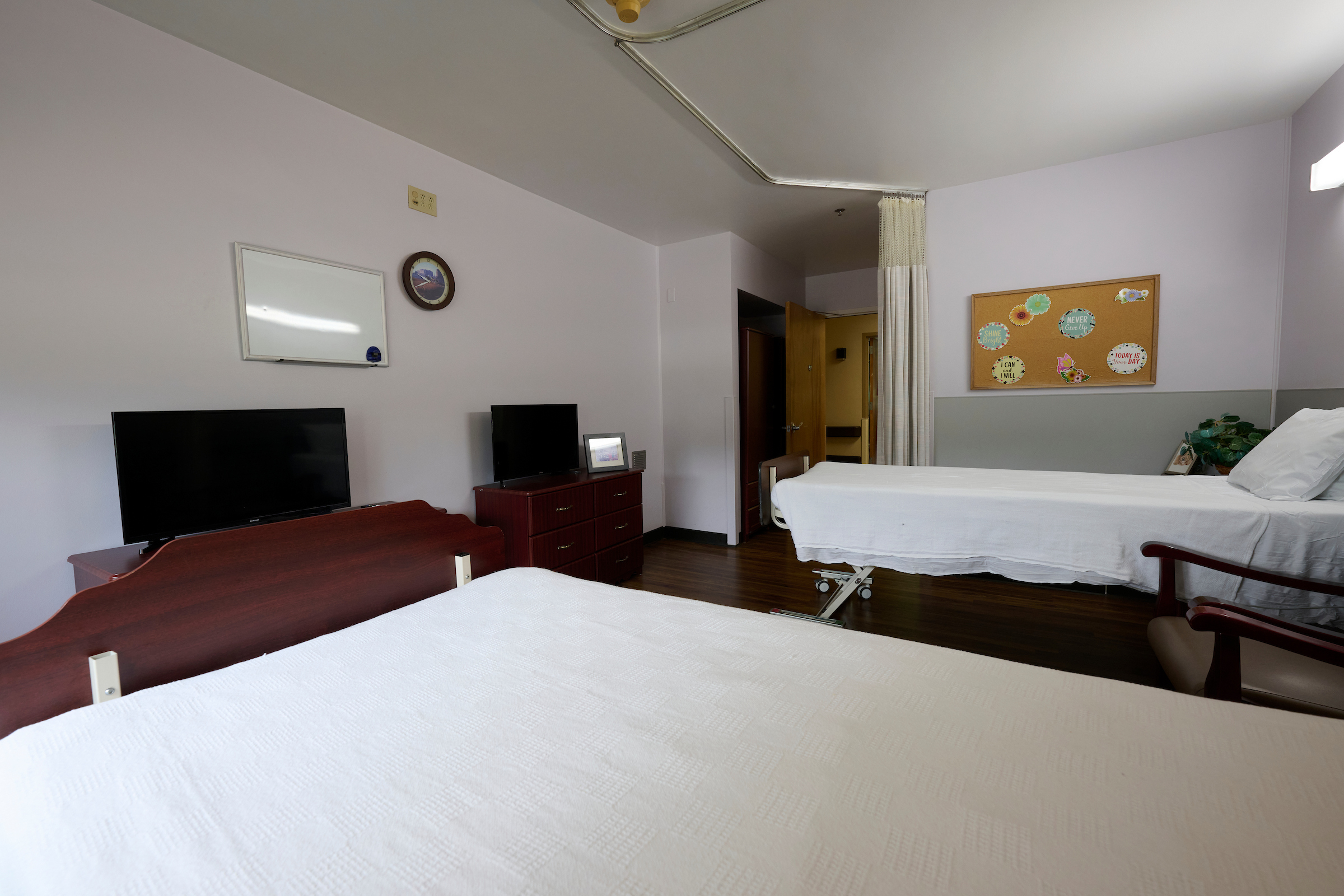 Hospital room with two beds, lavender walls, and a corkboard displaying motivational quotes on the right.