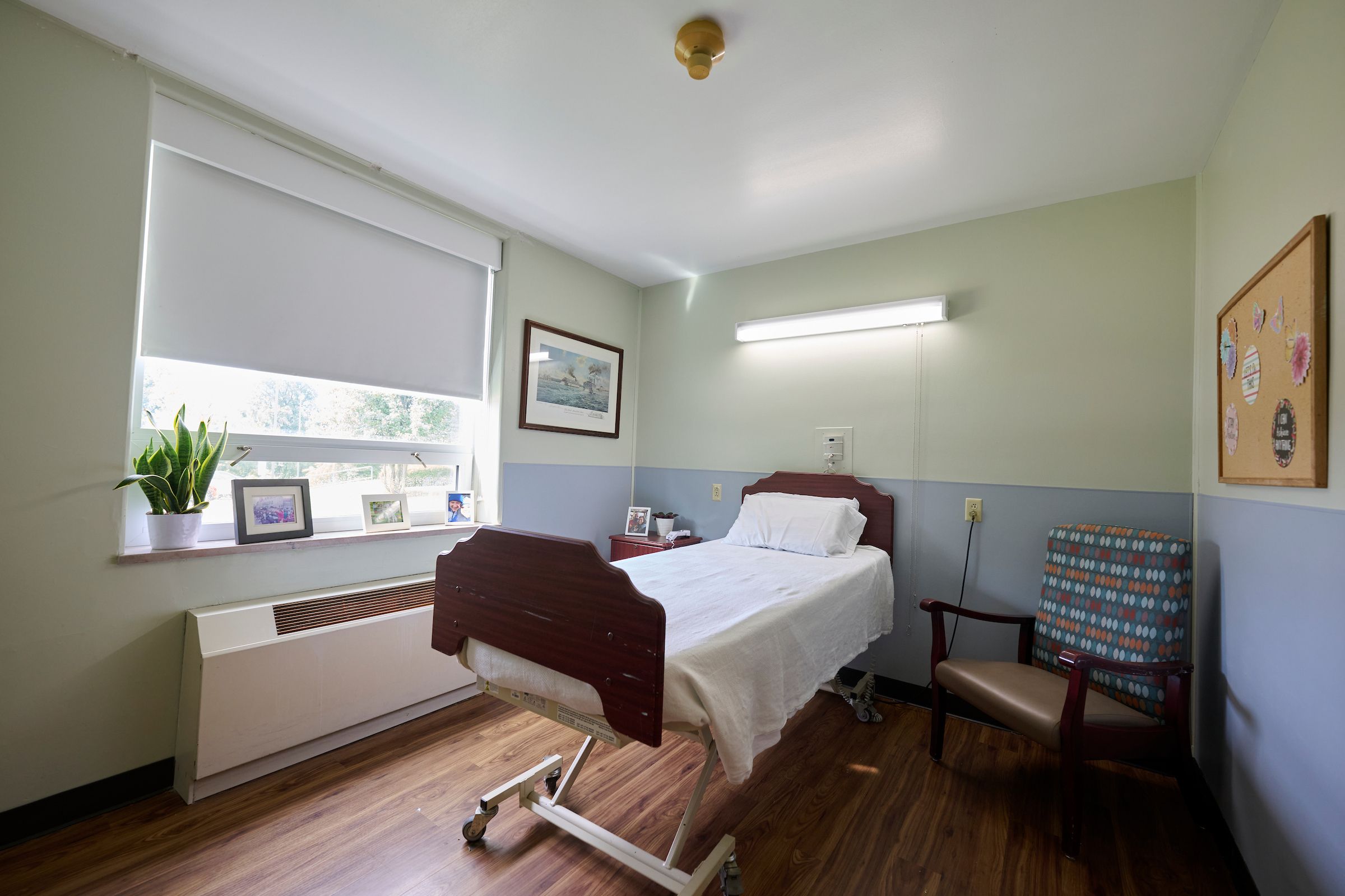 Hospital patient room with a single adjustable bed, white sheets, and a wooden headboard next to a window with closed blinds.