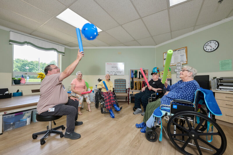 Older adults in a cheerful activity room playing with foam noodles and a blue polka-dot balloon, some in wheelchairs or chairs, smiling and engaging.