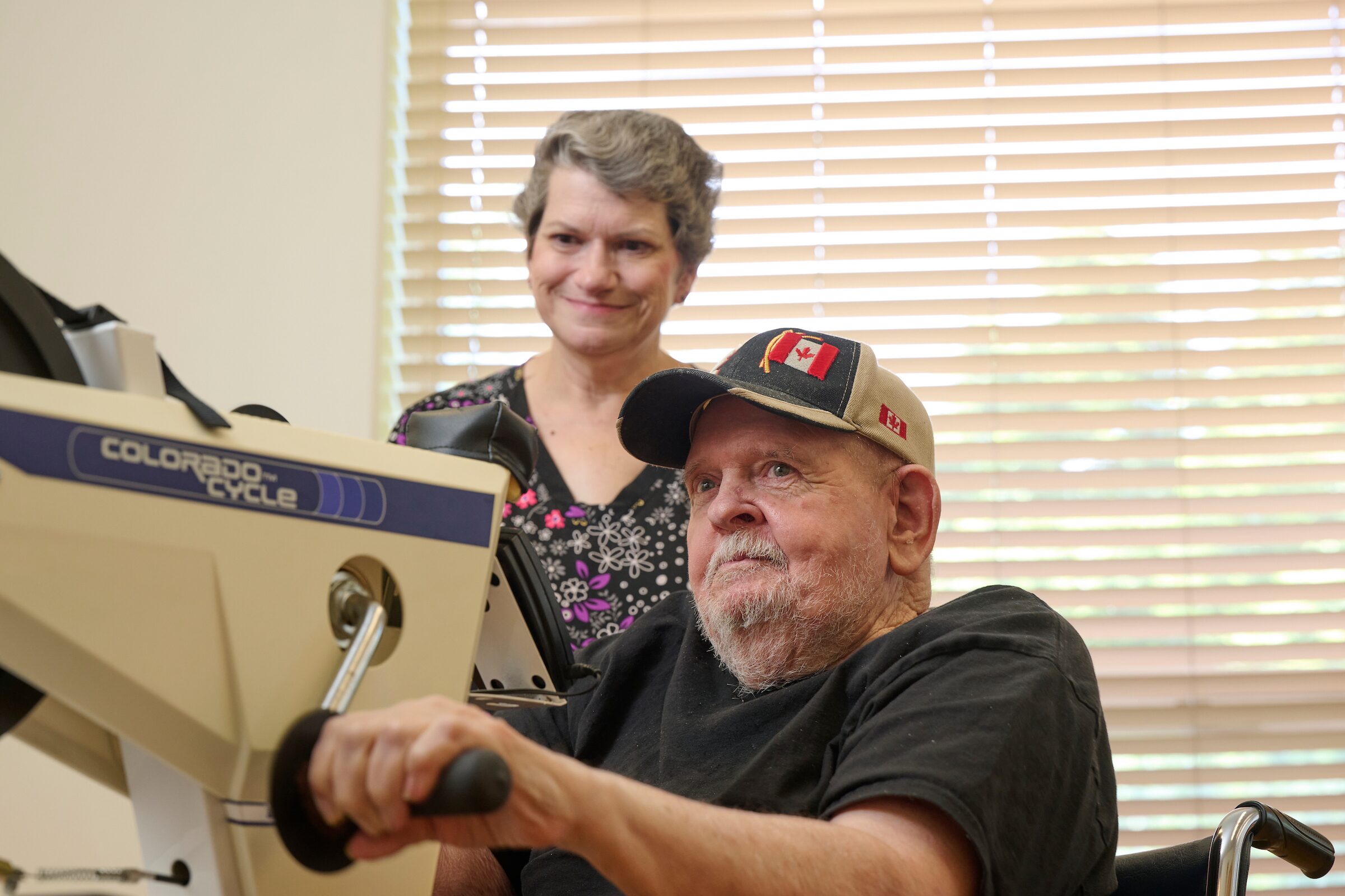 Older man operating a sewing machine with a smiling woman standing behind him in a sunlit room.