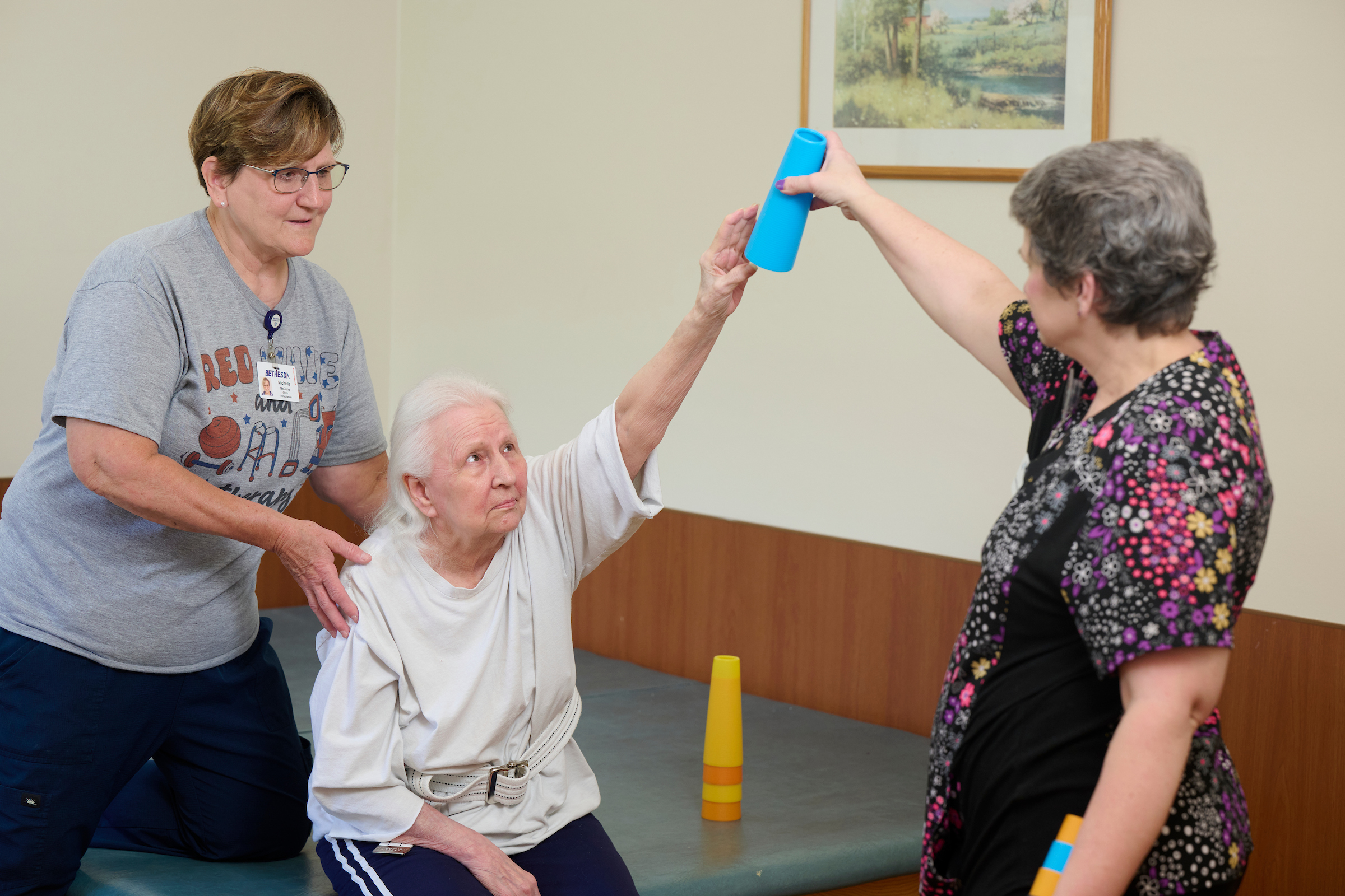Three women in a room share a blue plastic cup during a group activity; one elderly woman reaches to take the cup from another helper, with stacked orange cups on the floor nearby.