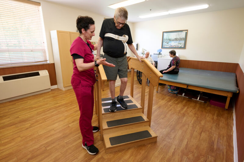 Nurse in pink scrubs assists an elderly man with a harnessed belt walking on parallel bars in a rehab room, other patients seated nearby.