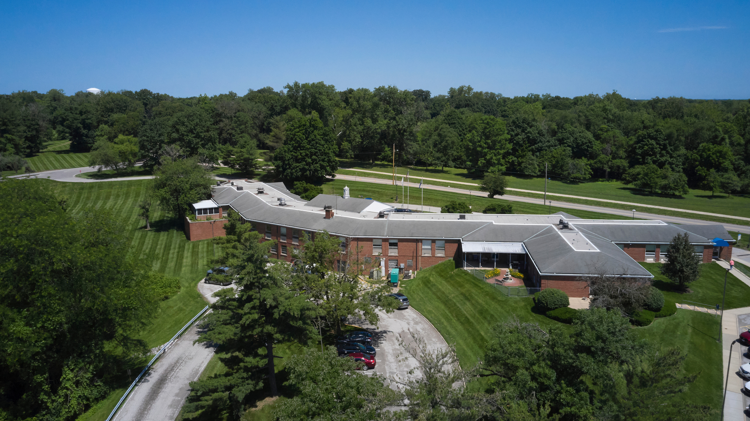 Aerial view of a brick campus building with gray roof surrounded by green lawns and trees under a blue sky.