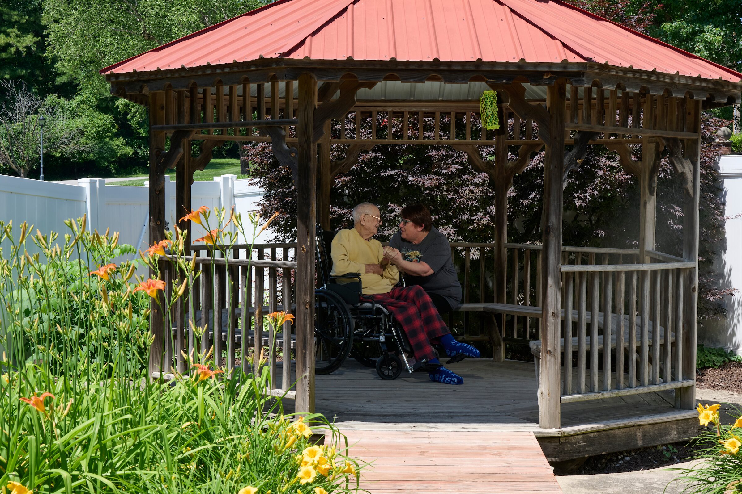 An elderly person in a wheelchair talking with a caregiver inside a wooden gazebo in a garden, with orange daylilies in the foreground and a red roof above.