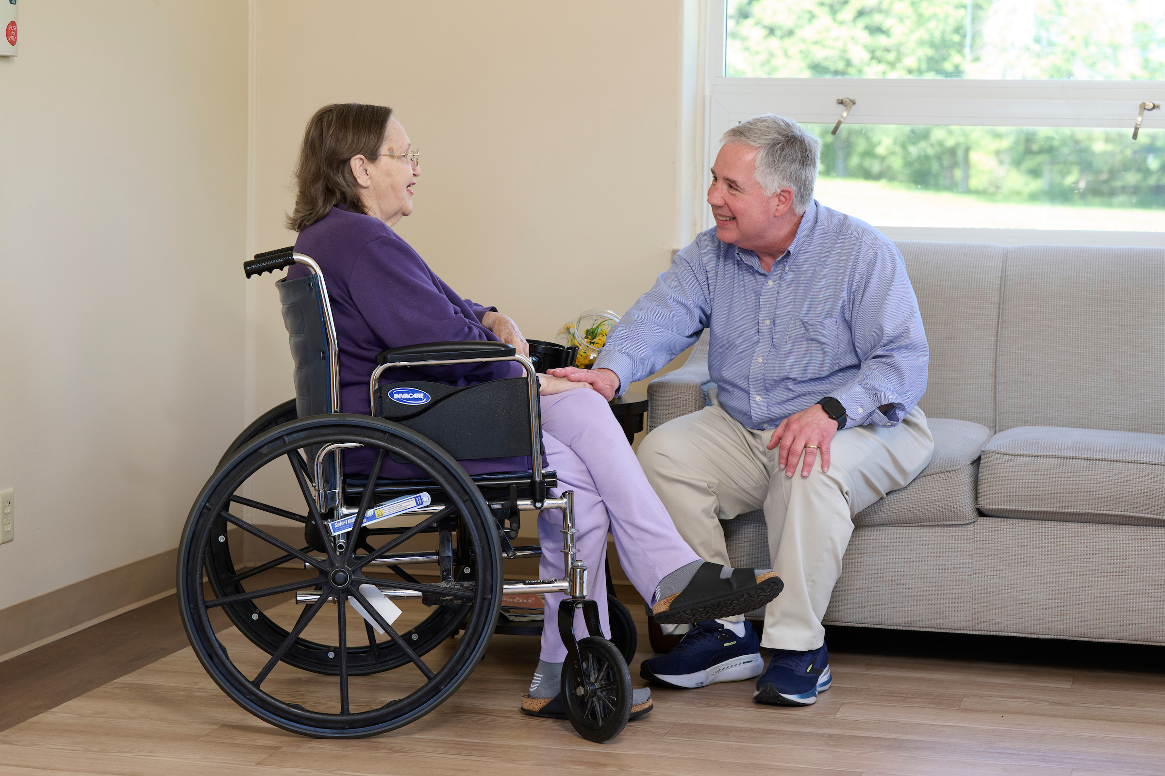 Elderly woman in a wheelchair holding hands and smiling with a man seated on a sofa in a bright, homey room close to a window.