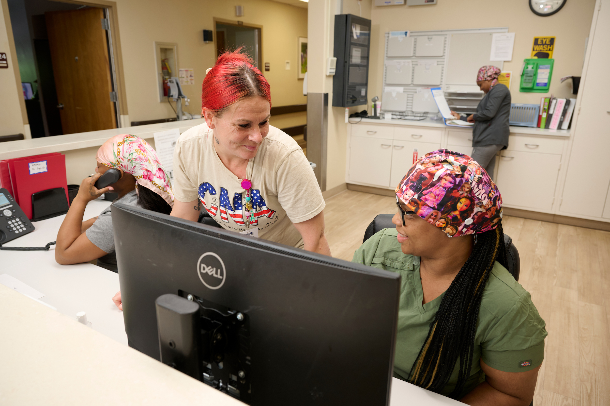 Three healthcare workers at a reception desk in a clinic; one woman with red hair leans toward a computer monitor while another patient with a colorful headscarf sits nearby, and a nurse sorts papers in the background.