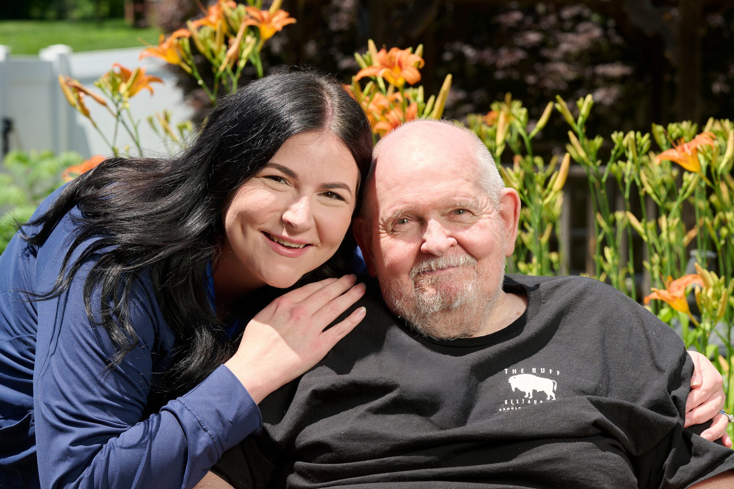 Woman with long dark hair smiles beside an older man with a beard in a sunny garden with orange daylilies behind them.