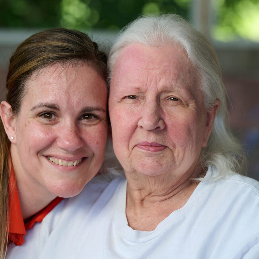 Two women embracing and smiling at the camera outdoors, one younger with brown hair on the left and one older with white hair on the right.