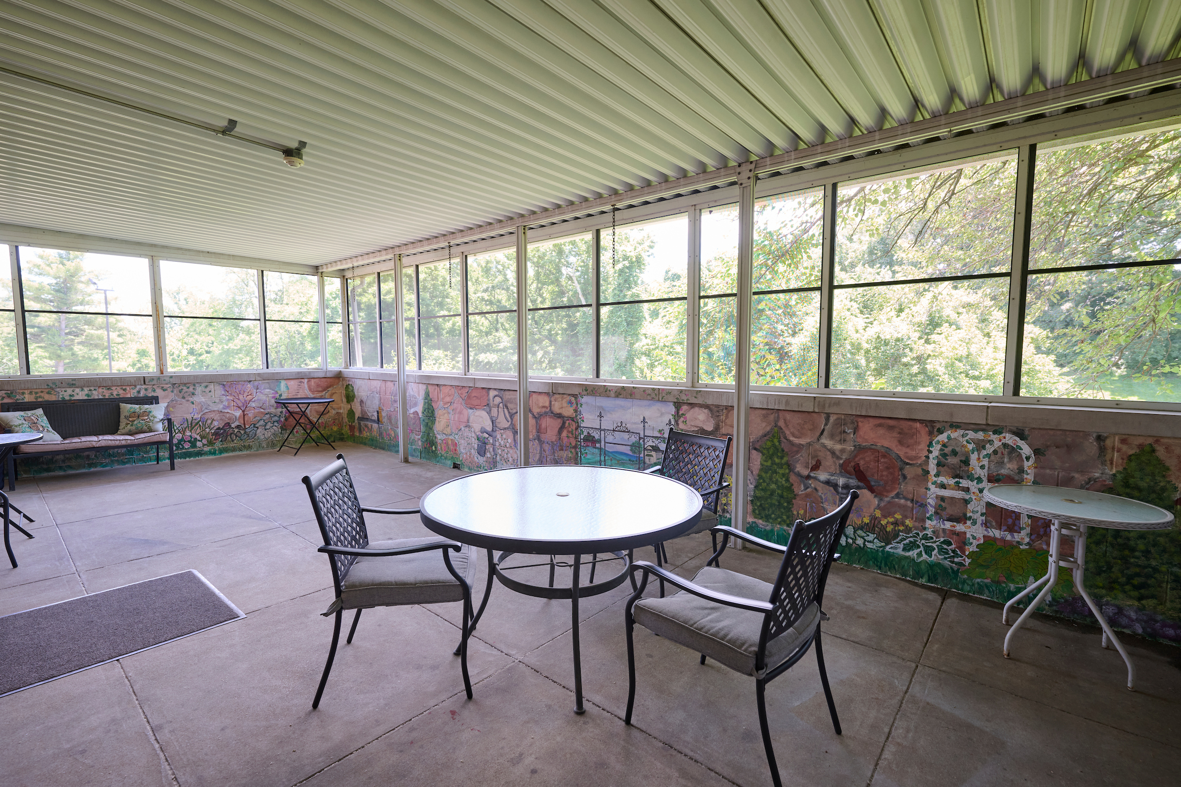 Enclosed patio with a round glass table and metal chairs, a colorful mural along the lower wall, and large windows showing trees outside.
