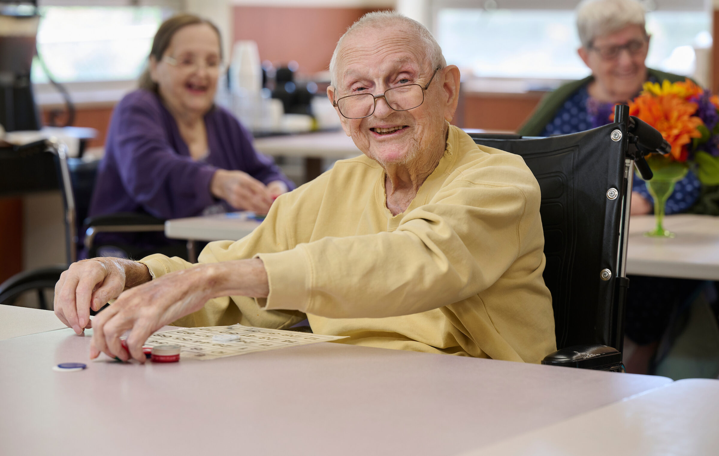 Smiling elderly man in a beige shirt in a wheelchair, reaching for bingo cards on a table.