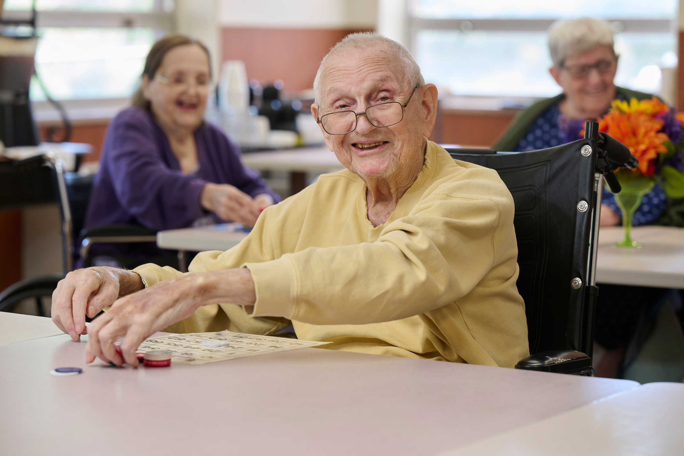 Smiling elderly man in a beige shirt in a wheelchair, reaching for bingo cards on a table.