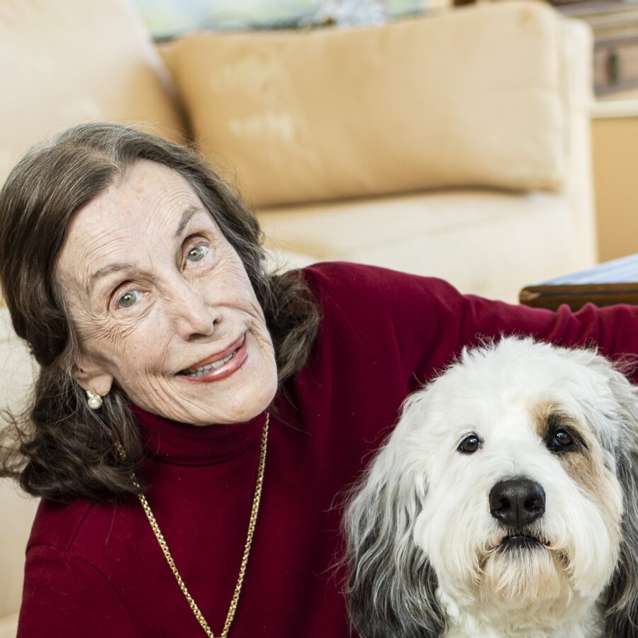 Smiling elderly woman in a burgundy sweater cuddling a fluffy white-and-gray dog on a sofa behind her.