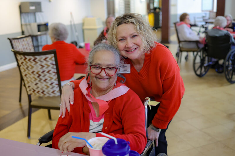 Two smiling elderly women in red sweaters pose together in a care facility's common area, one in a wheelchair in front.
