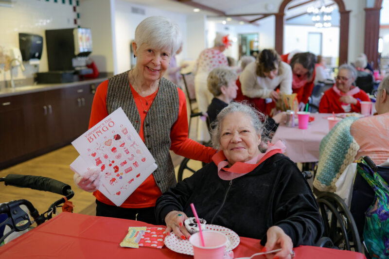 Smiling elderly woman standing with Valentine’s Day bingo cards while a senior in a wheelchair sits at a festively decorated table in the background.