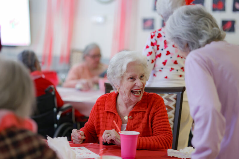 Joyful elderly woman in a red sweater laughing at a celebration table with pink cups nearby.