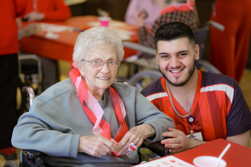 Smiling elderly woman in a wheelchair at a table with red decorations, assisted by a smiling staff member in a red uniform.