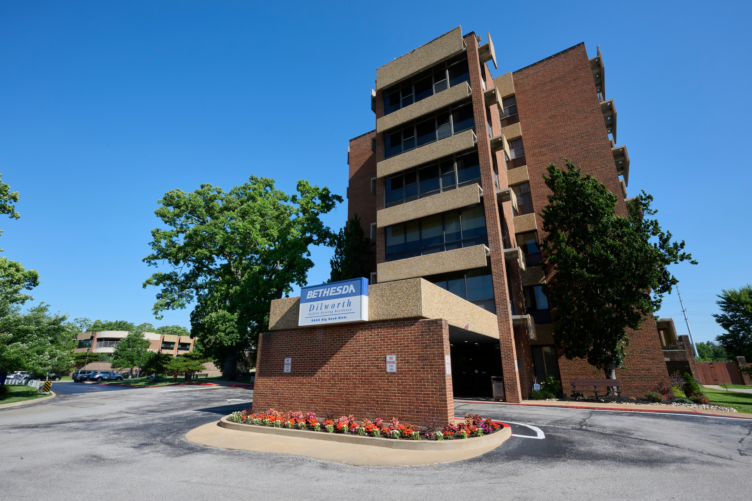 Five-story brick building with a Bethesda Dilworth sign, circular driveway, and a flower bed at the entrance.