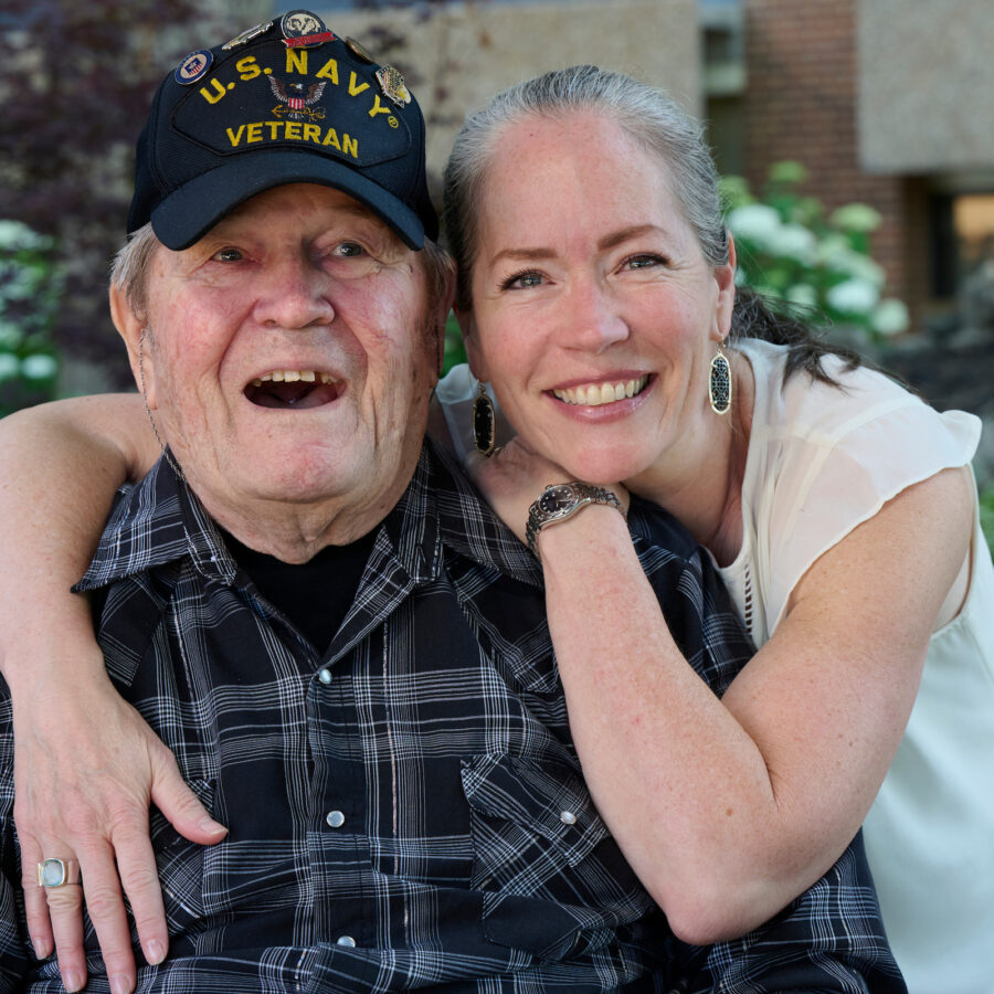 Elderly man wearing a U.S. Navy Veteran cap smiles as a woman hugs him from the side outdoors, both looking happy.