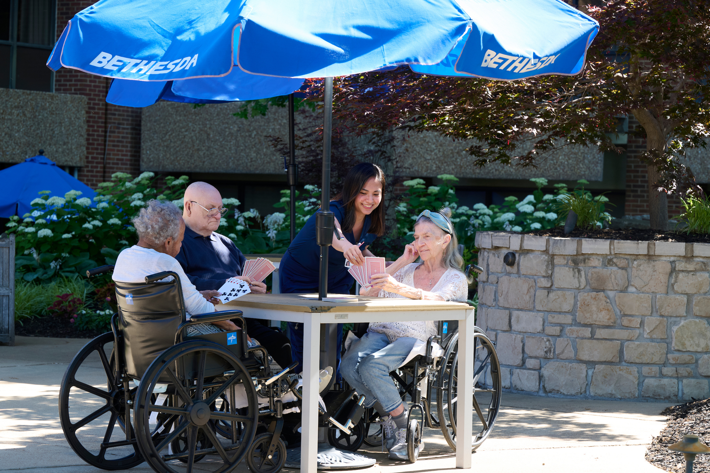 Seniors in wheelchairs around a table outdoors, playing cards with a caregiver assisting under a large blue umbrella.