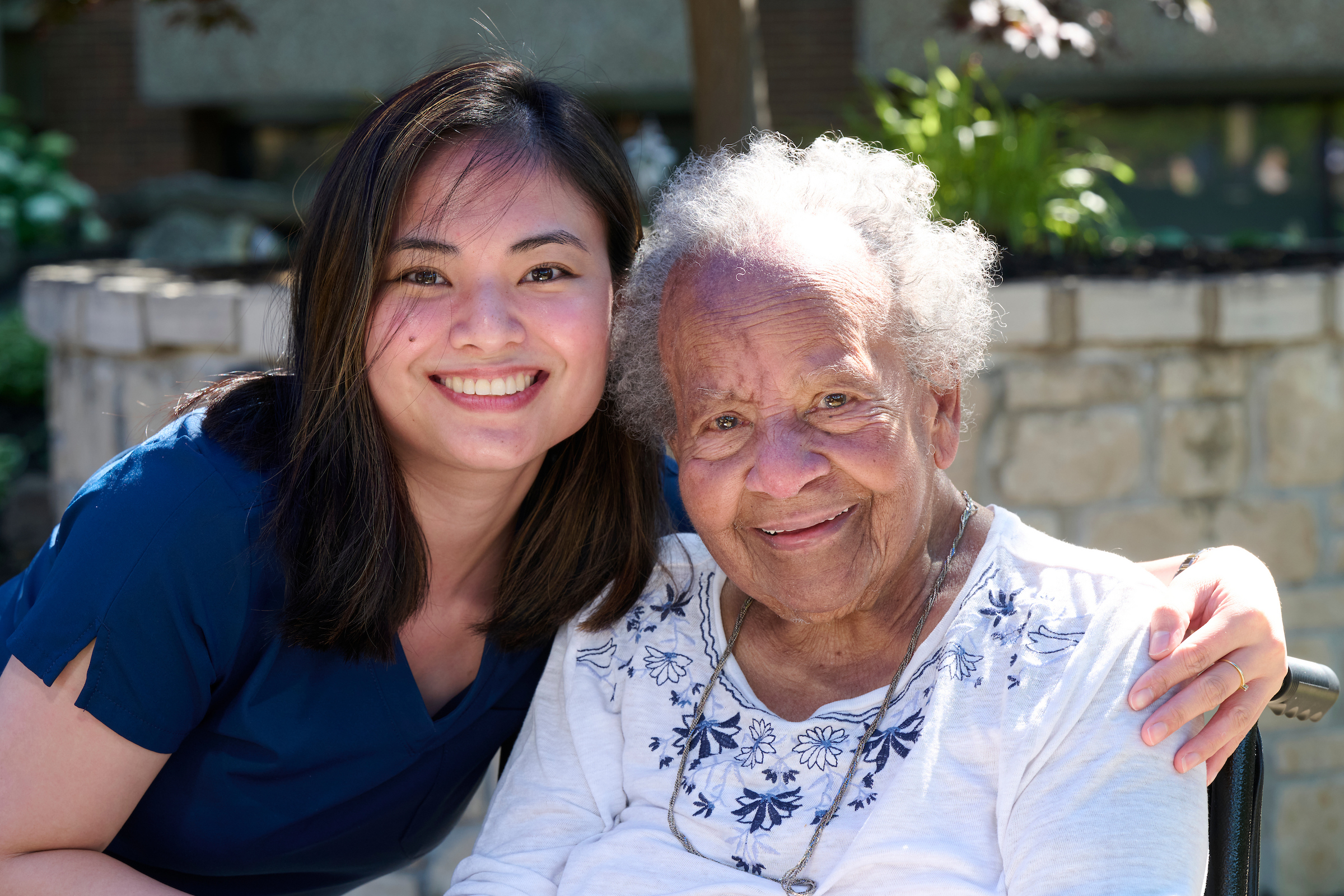 Two women smiling together outdoors, a younger woman on the left and an older woman on the right, near a stone wall and greenery.