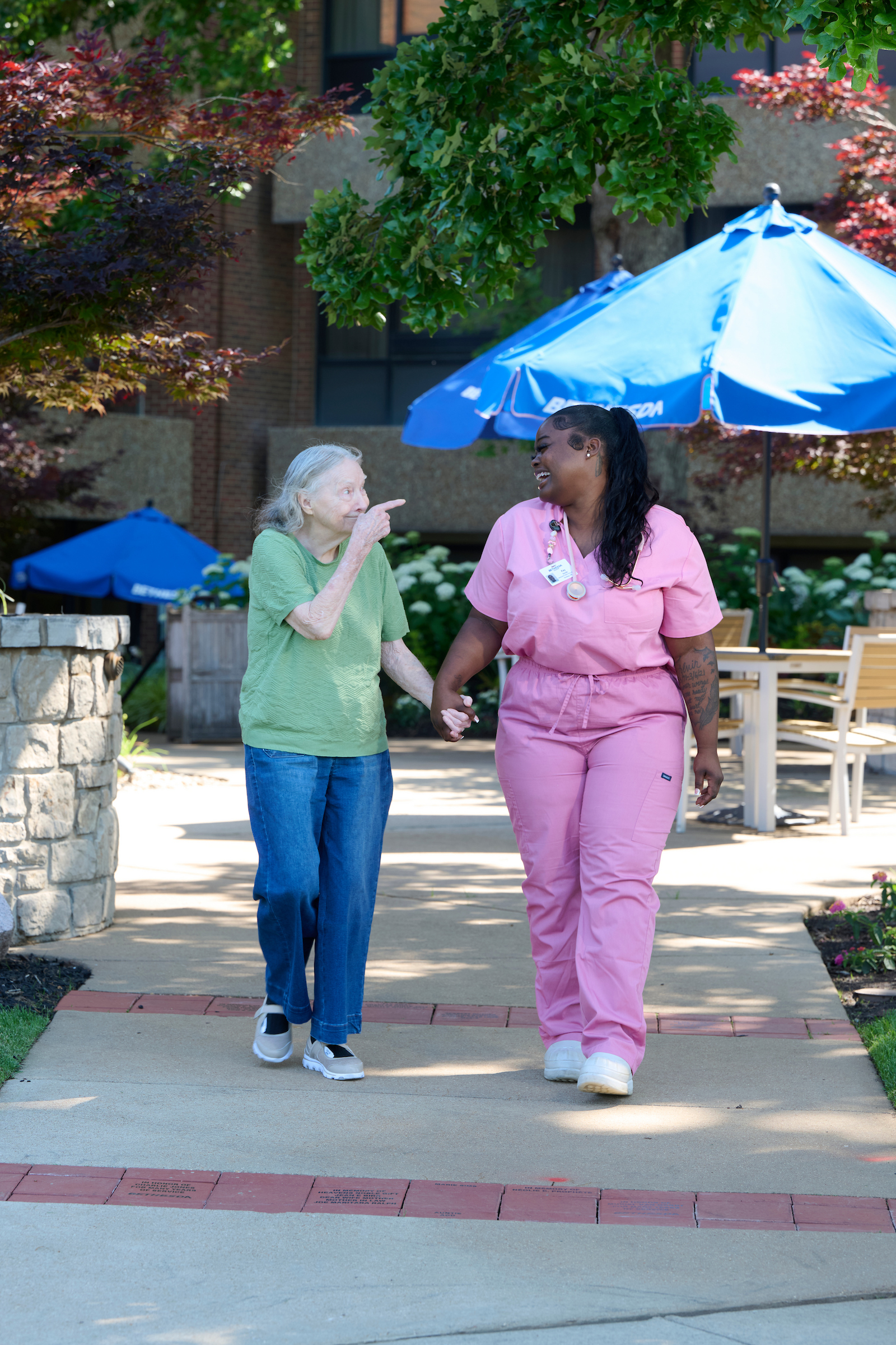 An elderly woman in a green shirt and jeans walking hand-in-hand with a healthcare worker in pink scrubs, smiling outdoors under blue umbrellas.