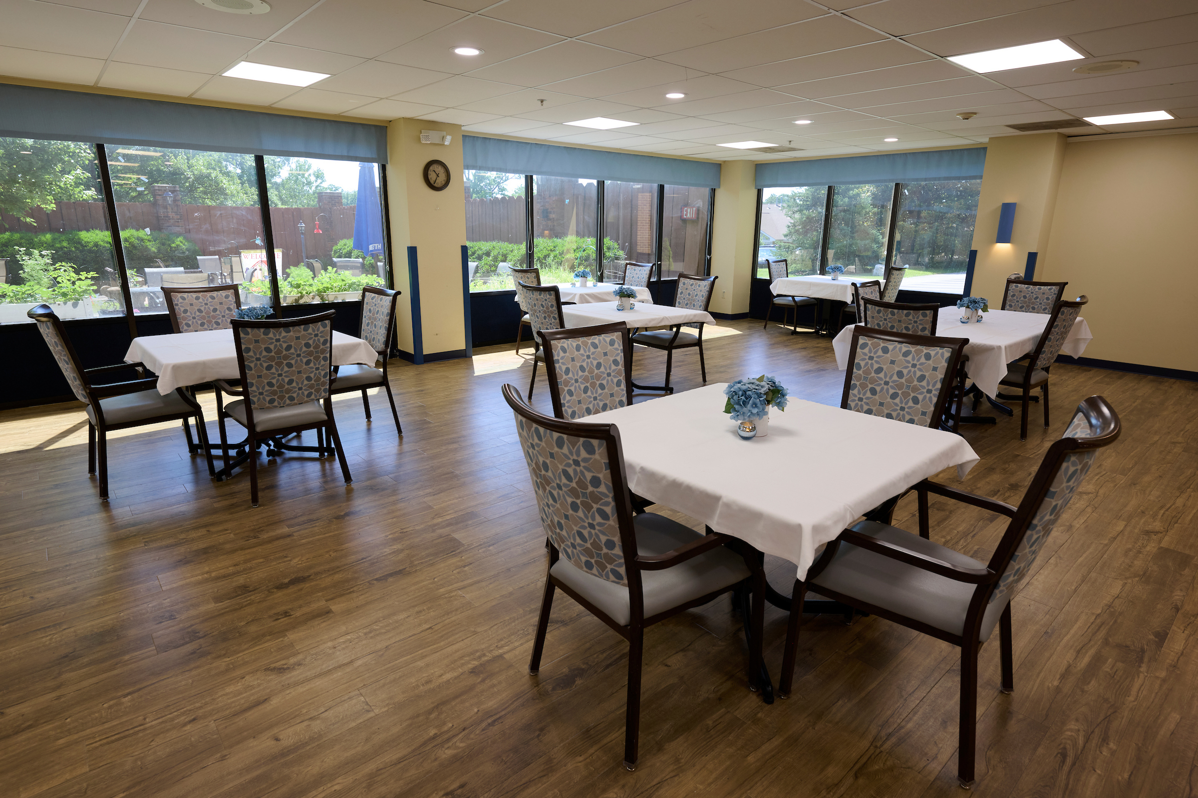 banquet-style dining room with round tables, white tablecloths, blue hydrangea centerpieces, and patterned chairs by large windows.