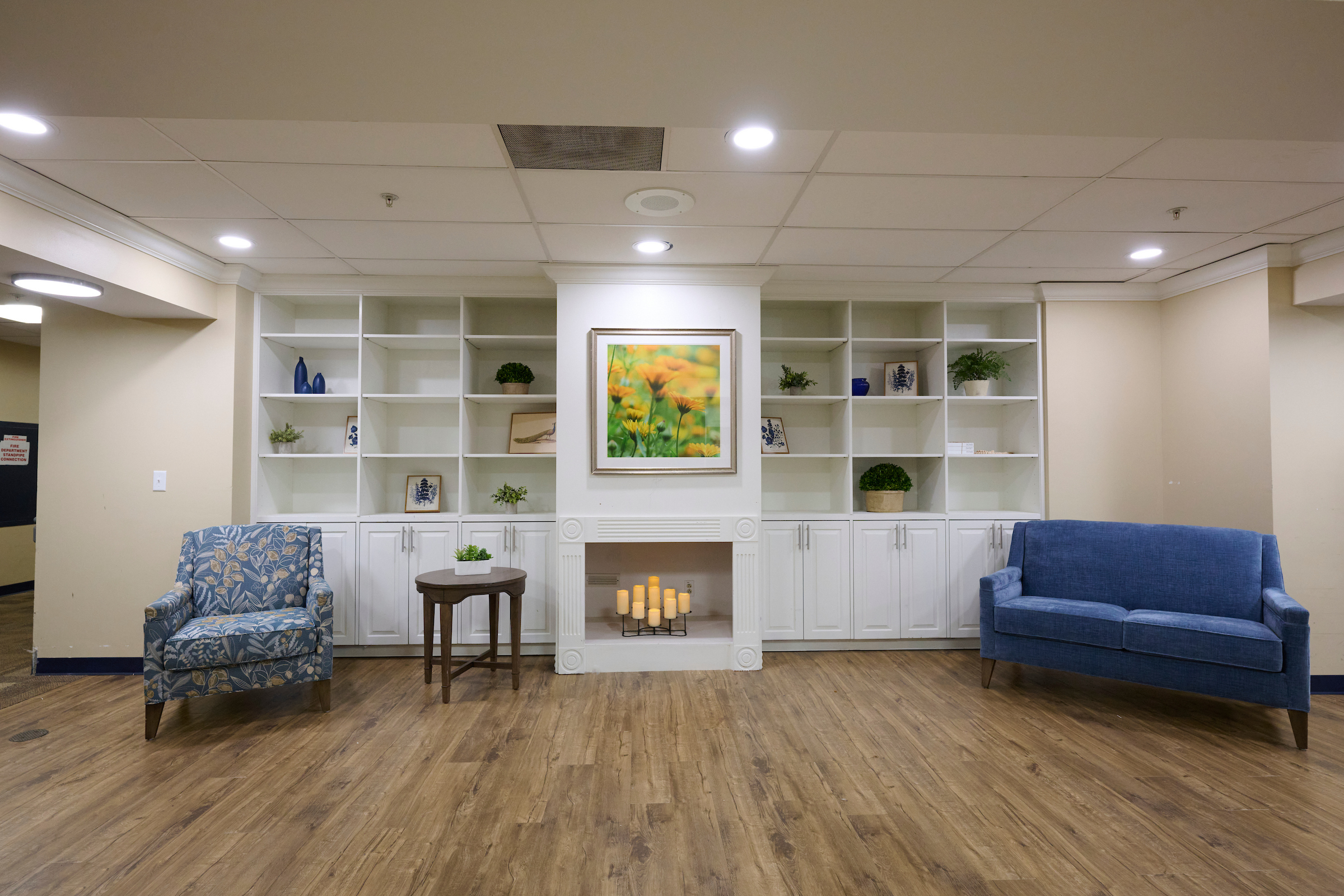 Living room with white built‑in shelves, a central fireplace, a framed floral painting, and blue armchairs on a wood floor.