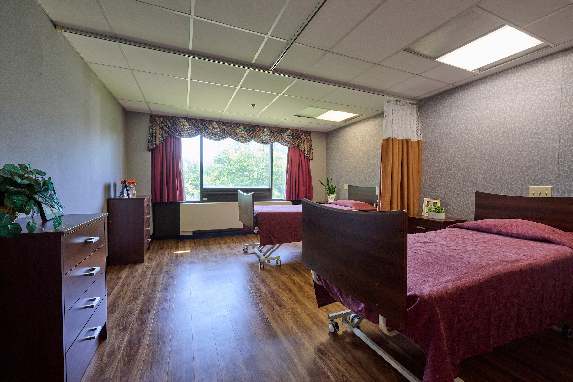 Hospital patient room with two burgundy bed covers, wooden furniture, and a large window with decorative red curtains nearby a desk and potted plant.