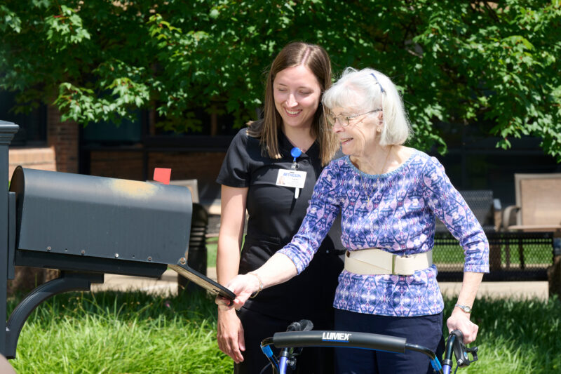 Caregiver in black scrubs helps an elderly woman adjust a blue Lumex bike outdoors, both smiling with green trees behind them.