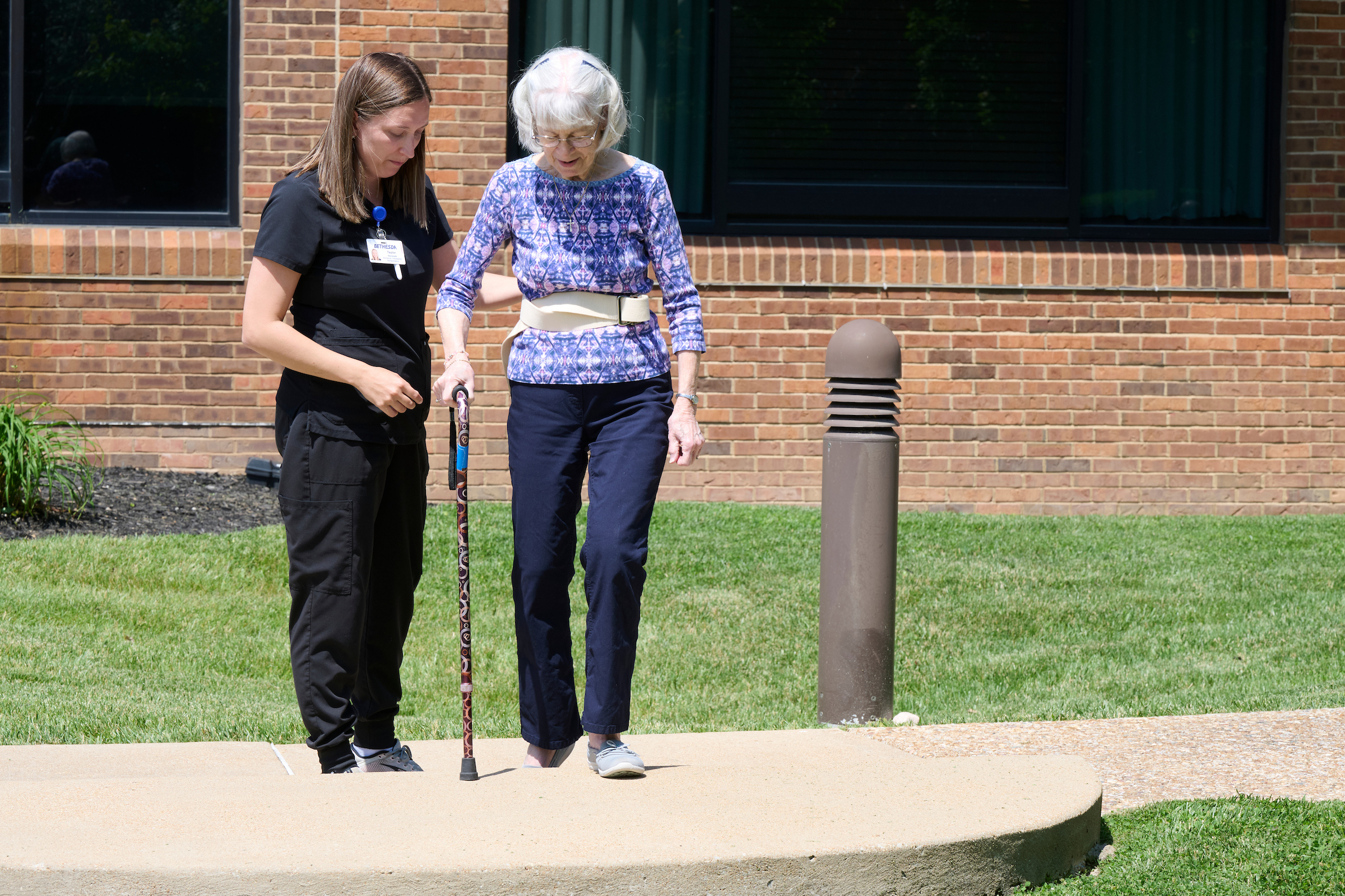Caregiver in black scrubs assists an elderly woman with a patterned cane outside a brick building.