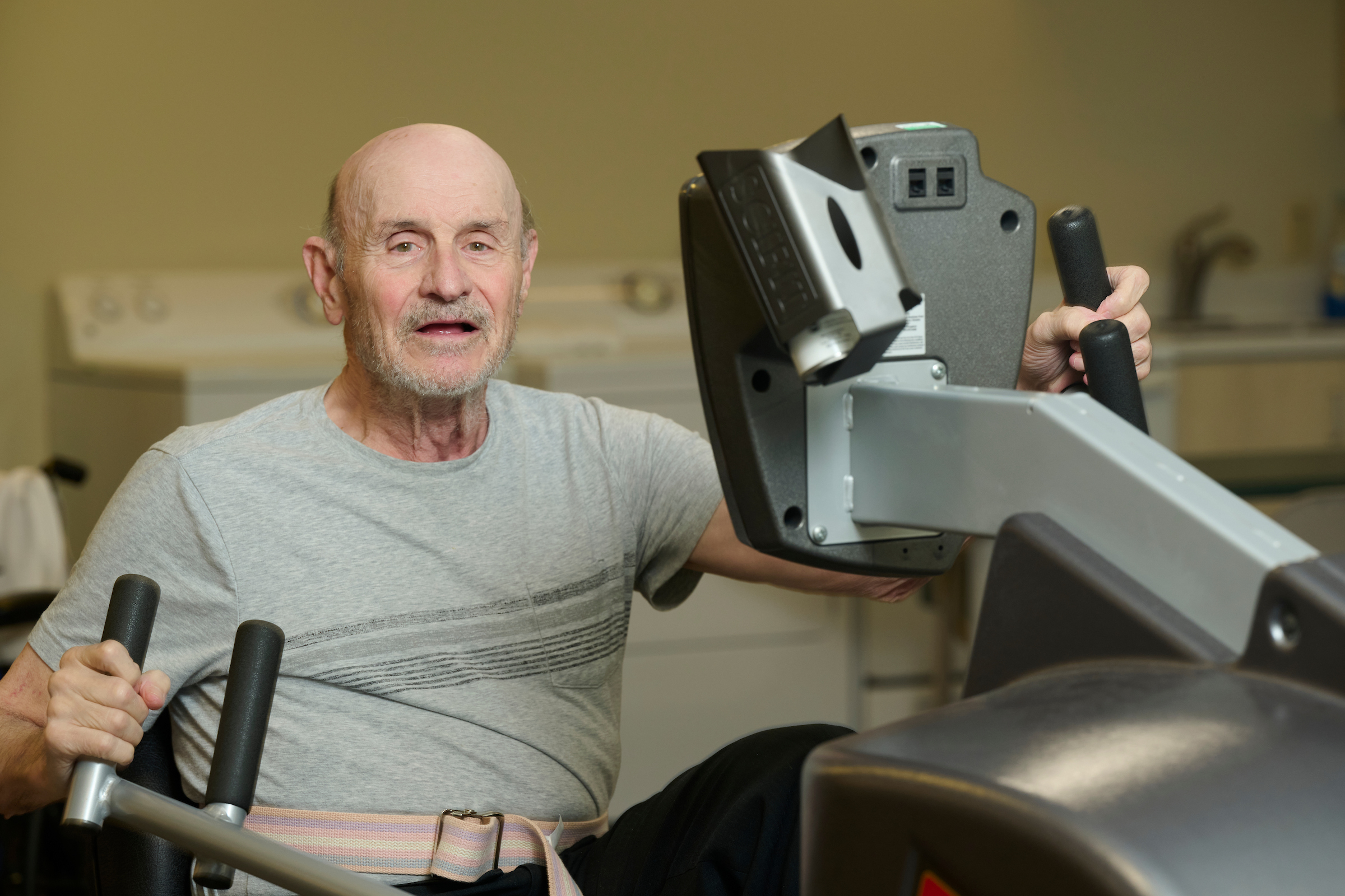 Older man in a gray t-shirt gripping the handlebars of a weight machine in a gym gym setting.