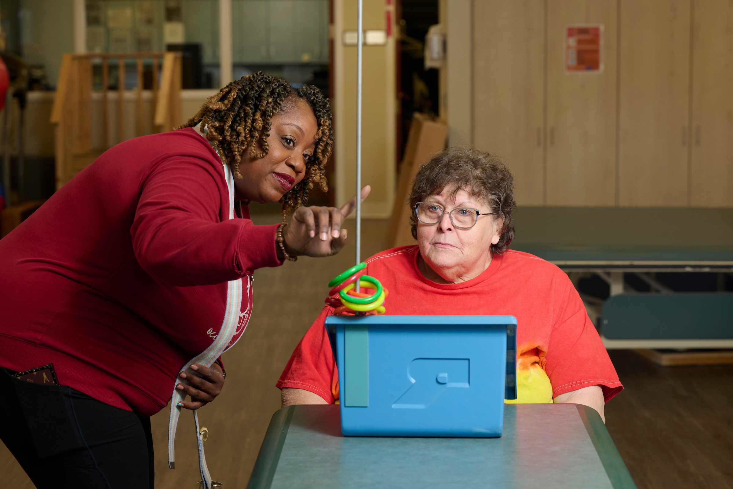 Caregiver leans toward a seated participant, guiding with a colorful spiral toy and a blue activity box on a table in a classroom setting.