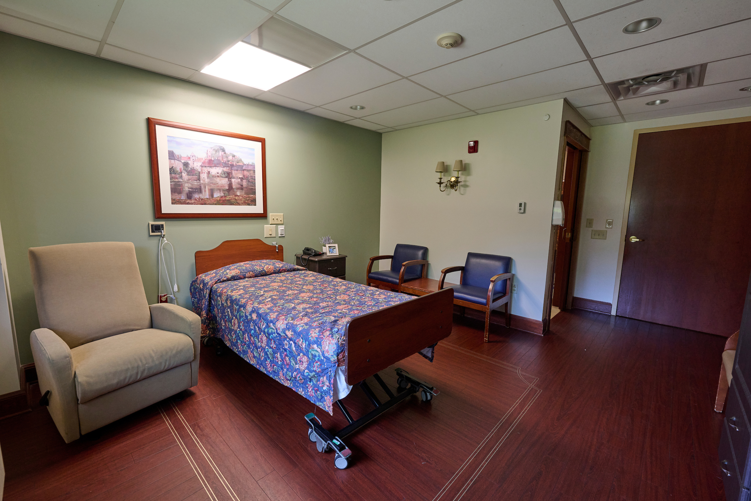 Patient room with a hospital bed on wheels, floral bedspread, beige armchair, and a framed landscape painting on a green wall.