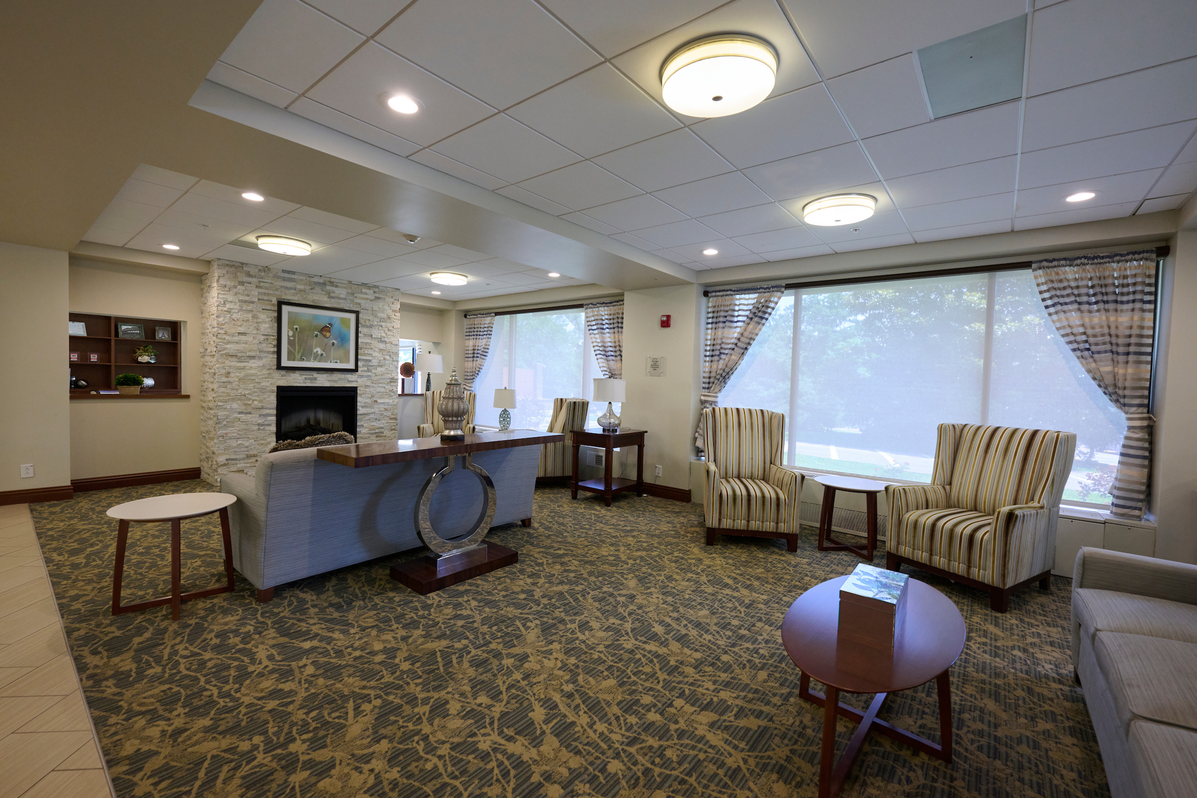 Hotel lounge with patterned carpet, beige seating, a stone fireplace, and large windows with striped curtains. Single round coffee tables sit nearby.