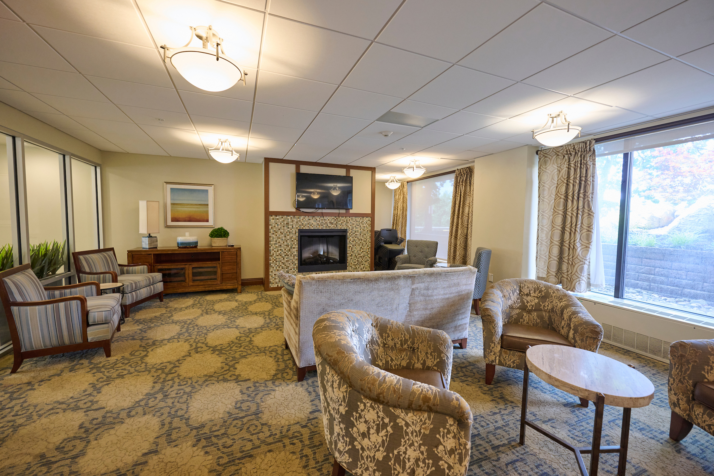 Cozy hotel-style living room with patterned armchairs around a fireplace and a wall-mounted TV above tiled surround, large windows on the right with curtains.